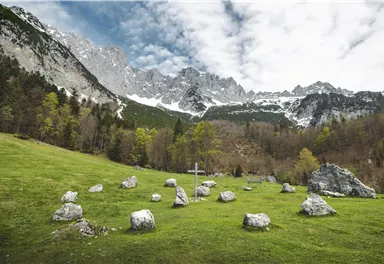 A green meadow with a stone circle and a wooden cross in the foreground. In the background, majestic mountains rise up under a cloudy sky.