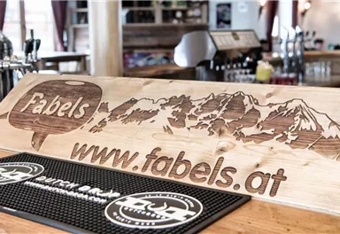 Eine Holztheke mit dem Schriftzug "Fabels" und einer Berglandschaft. Die Website www.fabels.at ist ebenfalls sichtbar.