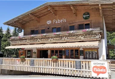 Ein schönes Holzgebäude mit einer einladenden Terrasse. Das Restaurant „Fabels“ hat ein rustikales Design und ist von Bäumen umgeben.