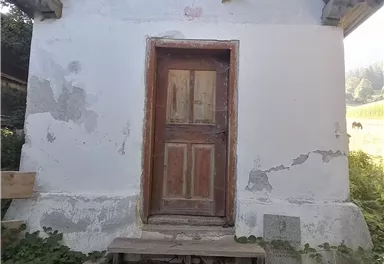 A small, simple building with a wooden front door. It stands in front of a green landscape and is surrounded by grass and plants.