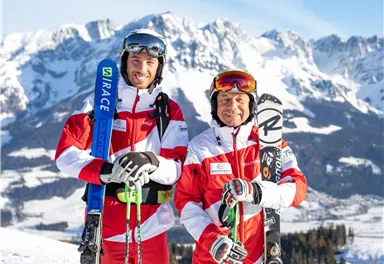Two skiers are standing on a snow-covered slope with majestic mountains in the background. They are wearing red and white ski clothing and are holding their skis in their hands.
