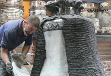 Ein Handwerker arbeitet an einer großen Glocke, die mit einer speziellen Mischung bearbeitet wird. Im Hintergrund sind weitere Glocken sichtbar.