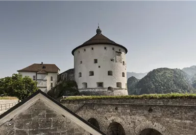Eine historische Festung auf einem Hügel, umgeben von grüner Landschaft und Bergen. Der Wachturm hat eine markante runde Form und verfügt über ein traditionelles Dach.