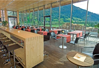 A modern restaurant with large windows and a view of the mountains. There is a long wooden bar and tables with red tablecloths.