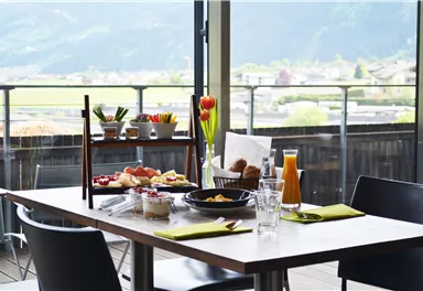 An elegant table with breakfast specialties and fresh flowers. In the background, the mountains and a green landscape are visible.