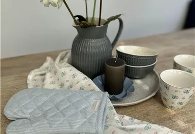 A stylish table setting with a gray pitcher, white flowers, and dishes. On the table, there is an oven mitt and a cloth.