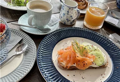 A delicious breakfast with an open sandwich topped with smoked salmon and cucumbers. Accompanied by coffee, juice, and some sweet treats on the table.