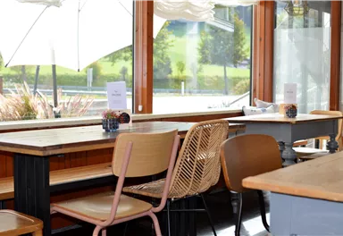 An inviting café with wooden chairs and tables. Large windows offer a view of the green surroundings.