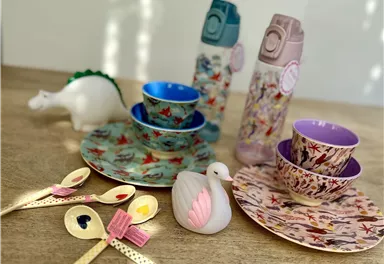 A colorful children's table with plates, cups, and spoons. Some bottles and a toy dinosaur are also visible.