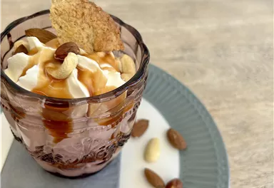 A dessert in a glass with whipped cream, caramel sauce, and nuts. Surrounding it are some almonds and cashew nuts.
