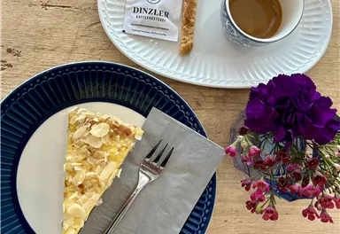 A beautiful plate with a slice of cake, garnished with almonds, and a fork. Next to it is a cup of coffee and a small sweet, surrounded by a colorful floral decoration.