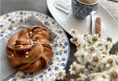 A sweet yeast knot on a blue plate with a cup of coffee beside it. In the background, there is a small vase with delicate flowers.