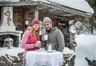 Ein Paar steht in der Winterlandschaft vor einer Hütte und hält Tassen. Die Umgebung ist verschneit und die Stimmung ist gemütlich.