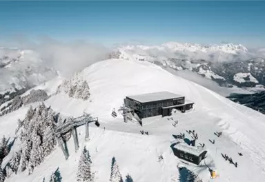 Ein schneebedeckter Berggipfel mit einer modernen Berghütte und Skiliften. Im Vordergrund sind einige Skifahrer und eine malerische Berglandschaft zu sehen.