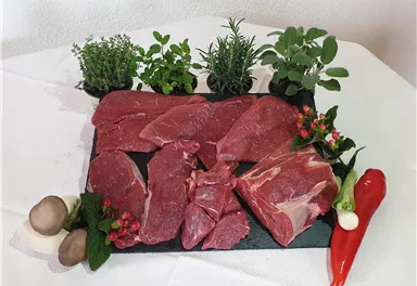 Frisches Fleisch auf einem schwarzen Steinbrett, umgeben von verschiedenen Kräutern wie Thymian und Rosmarin. Dazu sind rote und grüne Paprika sowie Champignons angeordnet.