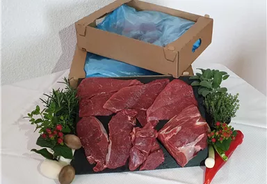 Frisches Fleisch auf einem schwarzen Granitbrett, umgeben von Kräutern und Gemüse. Im Hintergrund steht eine offene Verpackung.