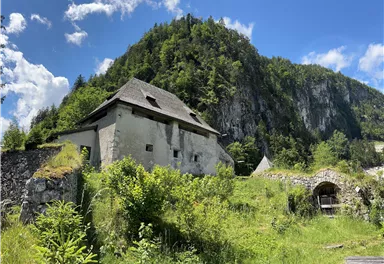 Eine alte, verfallene Gebäude inmitten von grüner Natur. Im Hintergrund ist ein hoher Berg mit Bäumen zu sehen.