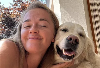 A woman smiles happily with a dog beside her.
They enjoy a sunny day outdoors.