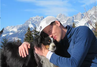 A man hugs his dog in the mountains. In the background, snow-covered mountains and a blue sky can be seen.