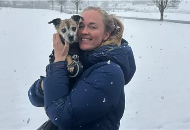 A smiling woman is holding a small dog in her arms. In the background, there is snow and it is lightly snowing.