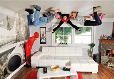 A creative room with an inverted perspective. Three people appear to be hanging from the ceiling, while a red sculpture man stands beside them.