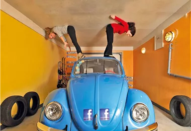 Two women are standing upside down on a blue beetle car in a garage. The walls are painted in a bright yellow, and tires can be seen in the background.