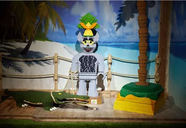 A colorful Lego figure with a pineapple on its head stands next to a Lego crocodile. In the background, there are palm trees and a blue sky.