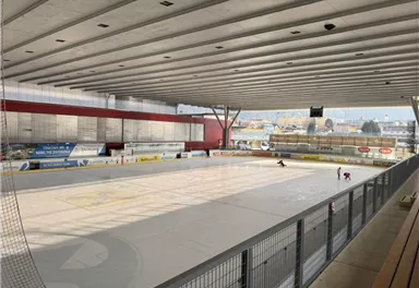 An ice rink with an empty ice surface and high ceilings. In the background, a few skaters can be seen.
