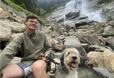 A young man is sitting on stones next to a dog and smiling. In the background, there is a waterfall and wooded hills visible.