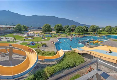 Ein Freizeitbad mit mehreren Schwimmbecken und Wasserrutschen. Im Hintergrund sind grüne Wiesen und Berge zu sehen.