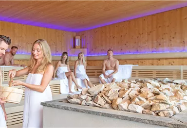 Ein entspannender Saunabereich mit Holzverkleidung. Zwei Personen bereiten sich auf das Saunaerlebnis vor, während andere Gäste entspannen.