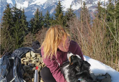 A man is sitting on a path and petting his dog. In the background, there are high mountains and a green valley.