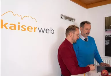 Two men are standing at a table and looking at a laptop. In the background, the logo of kaiserweb can be seen.