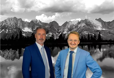 Two men in suits are standing in front of an impressive mountain landscape. The background shows the mountains and a calm lake, while the sky is cloudy.