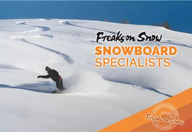 A snowboarder is riding through fresh snow in the mountains. In the background, clear blue skies and turns in the snow can be seen.