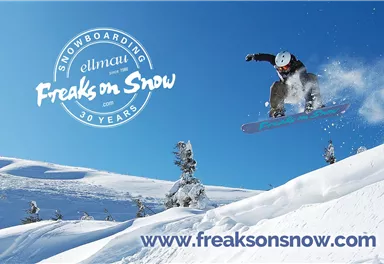 A snowboarder jumps over a snow-covered slope under a clear blue sky. Snow-capped mountains can be seen in the background.