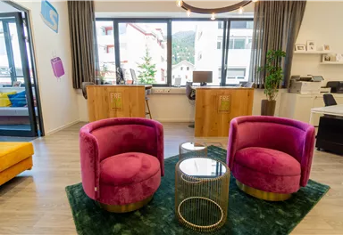 A modern office with two pink chairs and a small table. Large windows provide plenty of daylight and a view outside.