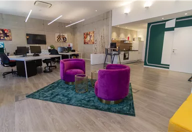A modern office with colorful chairs and a green carpet. There are several workstation areas and an open kitchen in the background.