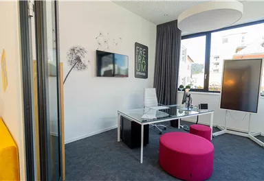 A modern office with a transparent desk and colorful seating options. Large windows provide plenty of daylight.