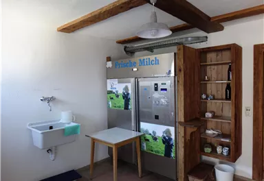 Eine gemütliche Küche mit einem Kühlschrank, der frische Milch anbietet. Ein Tisch und eine Holzregal ergänzen den Raum.