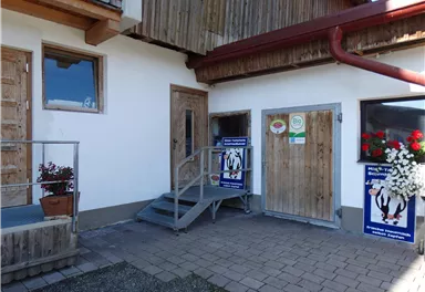 Ein Gebäude mit Holzverkleidung und zwei Türen. Vor der Eingangstreppe stehen Pflanzen und Plakate.