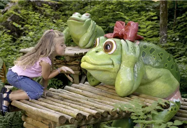 Ein Mädchen küsst eine große, grüne Kunststoffkröte mit einer roten Schleife. Im Hintergrund sind weitere Kröten und viel grünes Laub zu sehen.