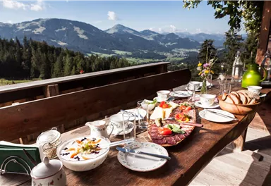 Ein schöner Tisch im Freien ist mit einer Vielzahl von Speisen und Getränken gedeckt. Im Hintergrund sind majestätische Berge und eine grüne Landschaft sichtbar.