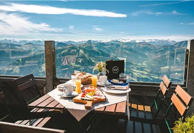 Ein gemütlicher Tisch mit Frühstück im Freien, umgeben von einer atemberaubenden Berglandschaft. Der Himmel ist klar und die Aussicht ist spektakulär.