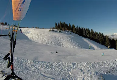 Eine schneebedeckte Berglandschaft mit klarem Himmel. Im Vordergrund ist ein Paragleiter mit einem bunten Gleitschirm zu sehen.