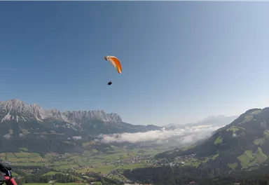 Ein Gleitschirmflieger schwebt über einer malerischen Berglandschaft. Das klare Wetter und die grünen Wiesen schaffen eine schöne Kulisse.