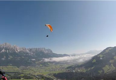 Ein Gleitschirmflieger schwebt über einer malerischen Berglandschaft. Das klare Wetter und die grünen Wiesen schaffen eine schöne Kulisse.