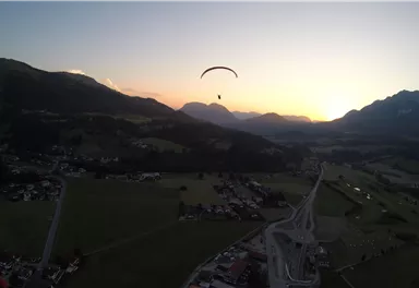 Ein Gleitschirmflieger schwebt über einer Landschaft mit Bergen und einem schönen Sonnenuntergang im Hintergrund. Die Umgebung ist ruhig mit grünen Wiesen und kleinen Dörfern.