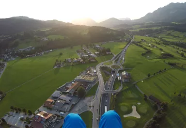 Eine atemberaubende Aussicht von oben auf eine grüne Landschaft mit einem kleinen Dorf und bergiger Kulisse. Die Sonne geht am Horizont unter und taucht die Szene in warmes Licht.
