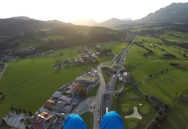 Eine atemberaubende Aussicht von oben auf eine grüne Landschaft mit einem kleinen Dorf und bergiger Kulisse. Die Sonne geht am Horizont unter und taucht die Szene in warmes Licht.